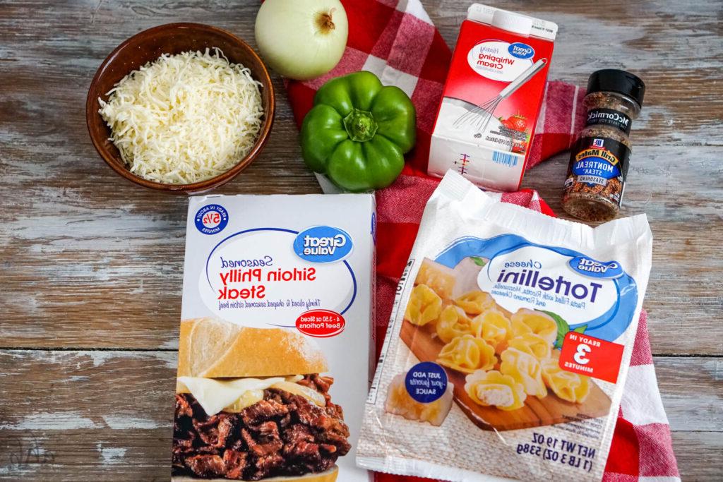 Montreal steak seasoning, heavy whipping cream, a green bell pepper, an onion, shredded provolone cheese, seasoned Sirloin Philly Steak, and a bag of frozen Cheese Tortellini on a red and white checked handkerchief.