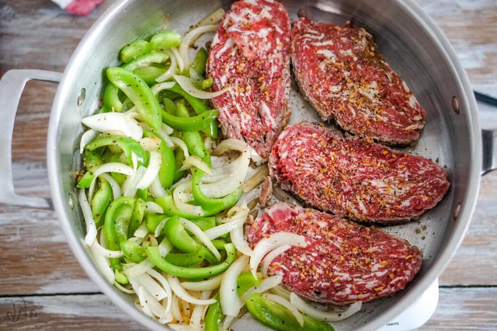 Four steaks cook in a sauté pan with slices of green pepper and onion.
